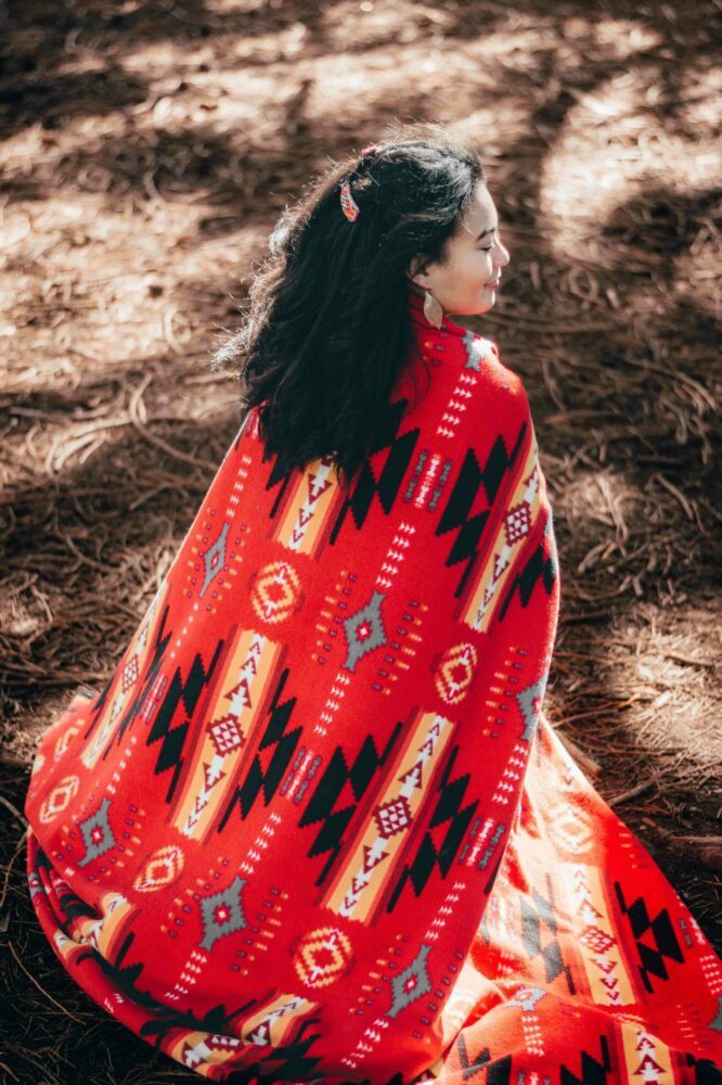An Indigenous woman kneels on the ground while being draped in a red shawl with Southwest Native American patterns
