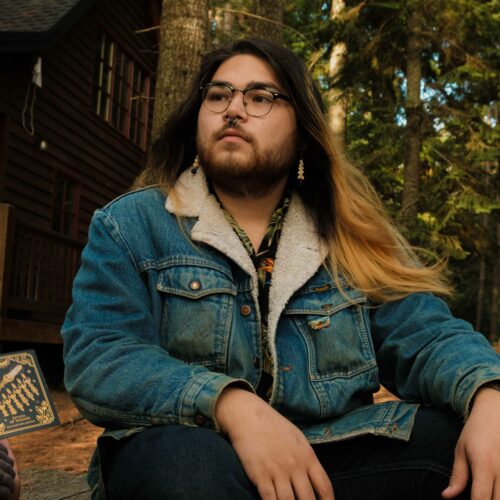 Kawika Maui Hā Kalama sits on a bench wearing a denim jacket with snail earrings staring at a tarot card in the forest